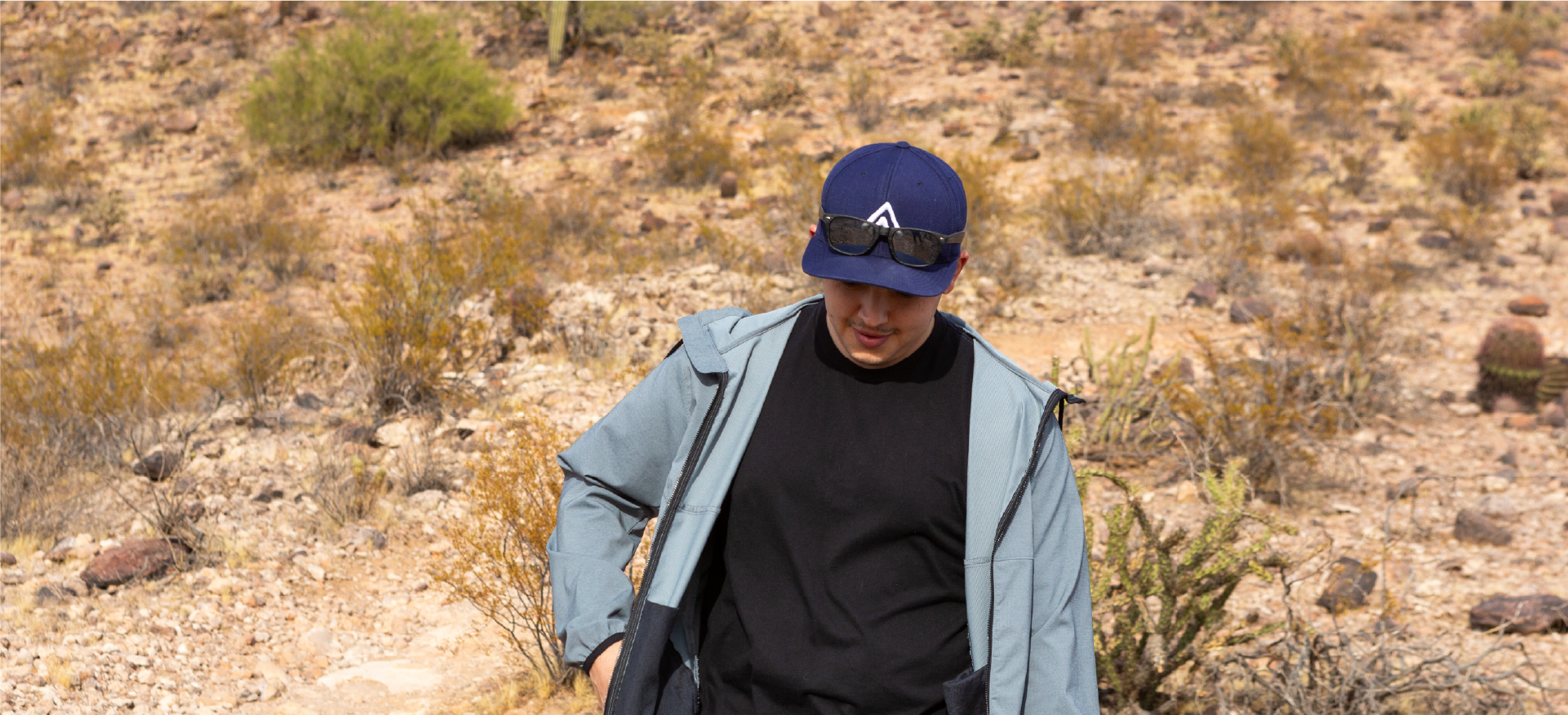 A man wearing a navy cap, sunglasses, black shirt, and a light blue jacket walking through a desert landscape with dry vegetation and rocky terrain in the background.