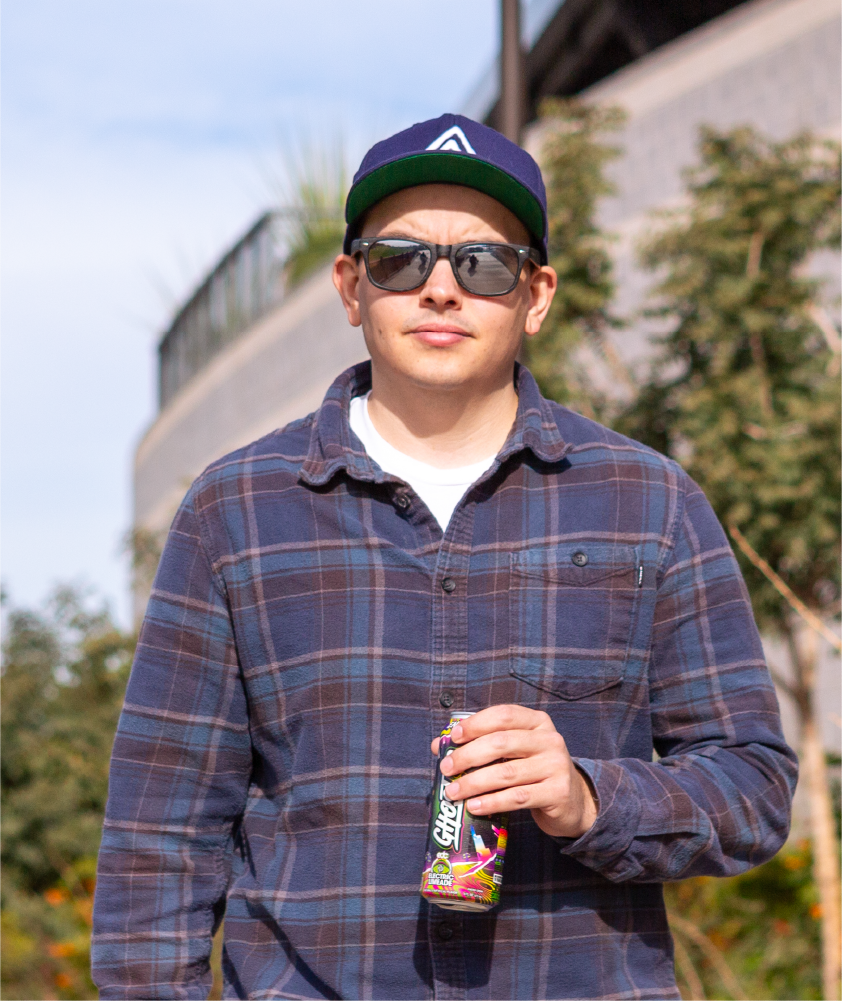 A man wearing a navy cap with a geometric logo, sunglasses, a plaid flannel shirt, and holding a colorful can of an energy drink, standing outdoors with trees and a modern building in the background.