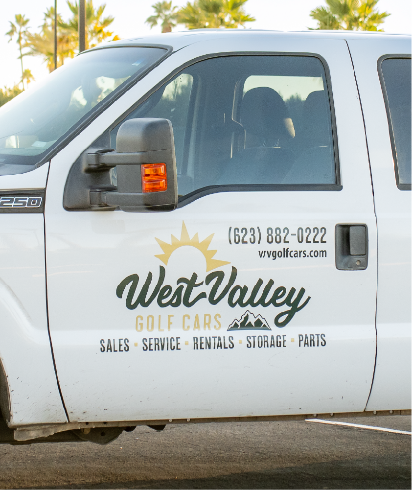 A white truck door displaying the West Valley Golf Cars logo, featuring a sun rising over mountains, along with text that includes the phone number '(623) 882-0222,' website 'wvgolfcars.com,' and services like sales, service, rentals, storage, and parts.