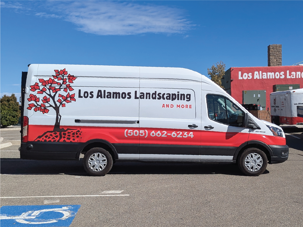 Right side view of Los Alamos Landscaping & More’s Transit van, displaying the company name, phone number, and a large red tree graphic extending from the back toward the front.